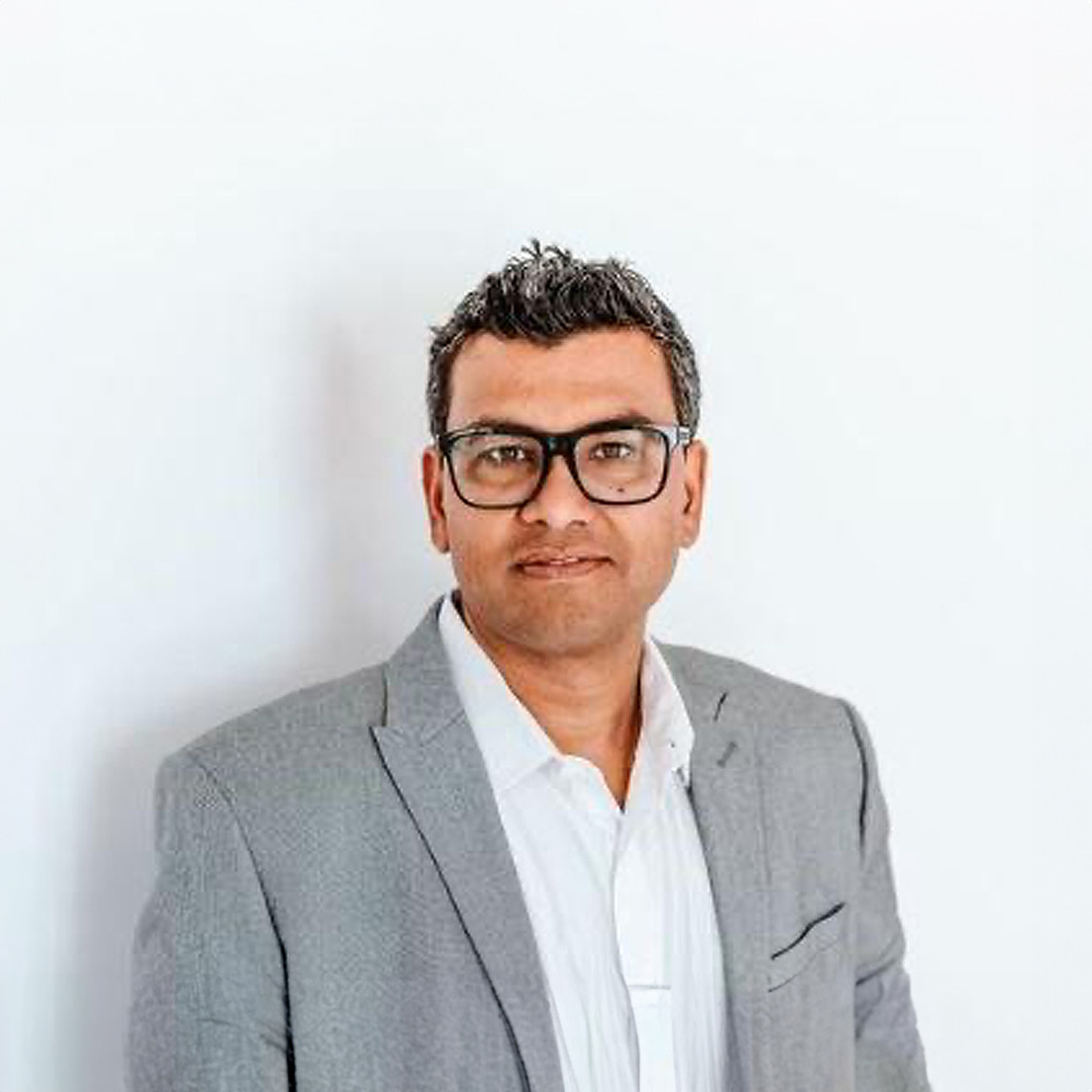 The image shows a man wearing glasses, a suit jacket, and a shirt standing against a light background with a neutral expression on his face.